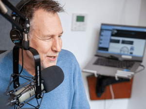 Kevin Roy speaks wears headphones and speaks into microphone, with laptop behind him