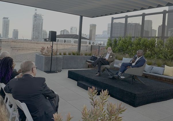 Audience members watch two speakers seated on a small stage during a rooftop panel discussion. A city skyline and landscaped terrace form the backdrop as one speaker talks into a handheld microphone.