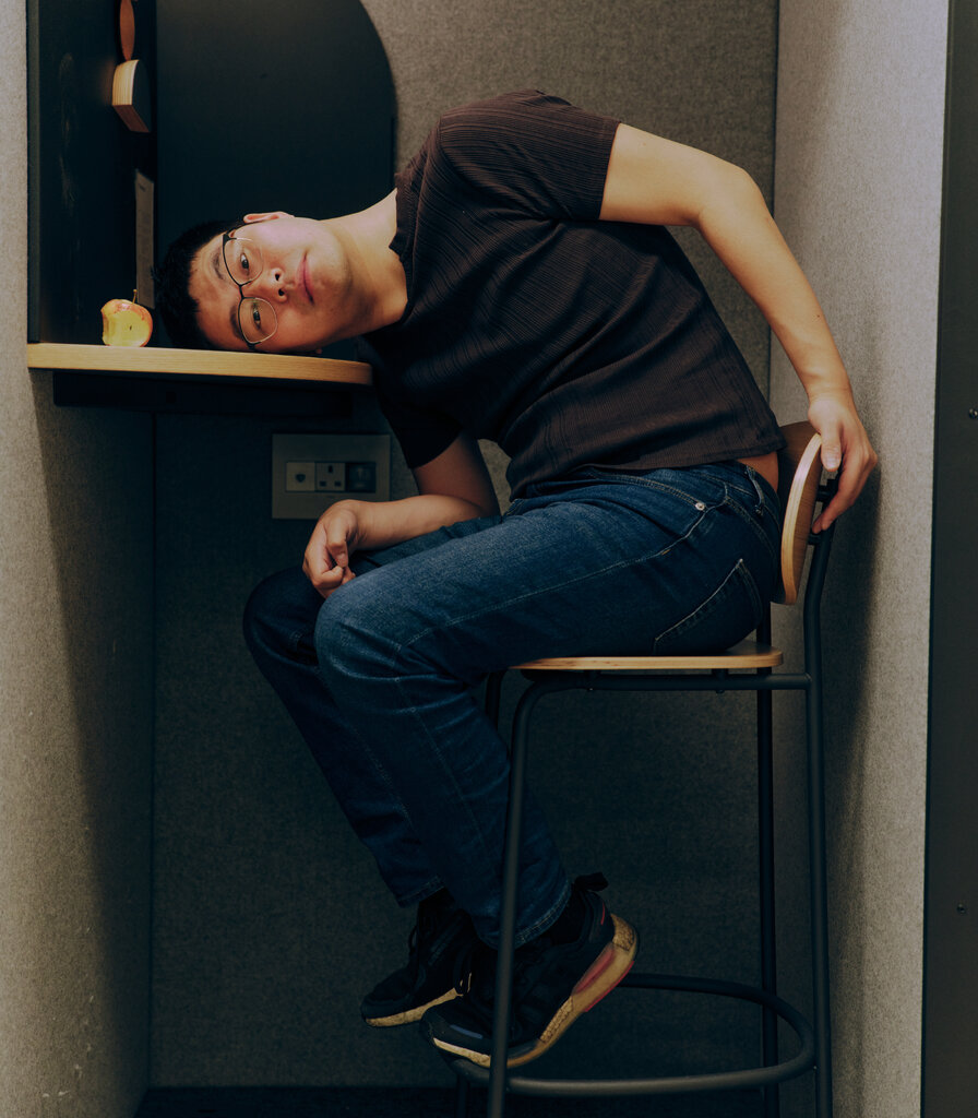 A man sits on a tall chair in a booth. His head leans on a desktop, were a bitten-apple sits.