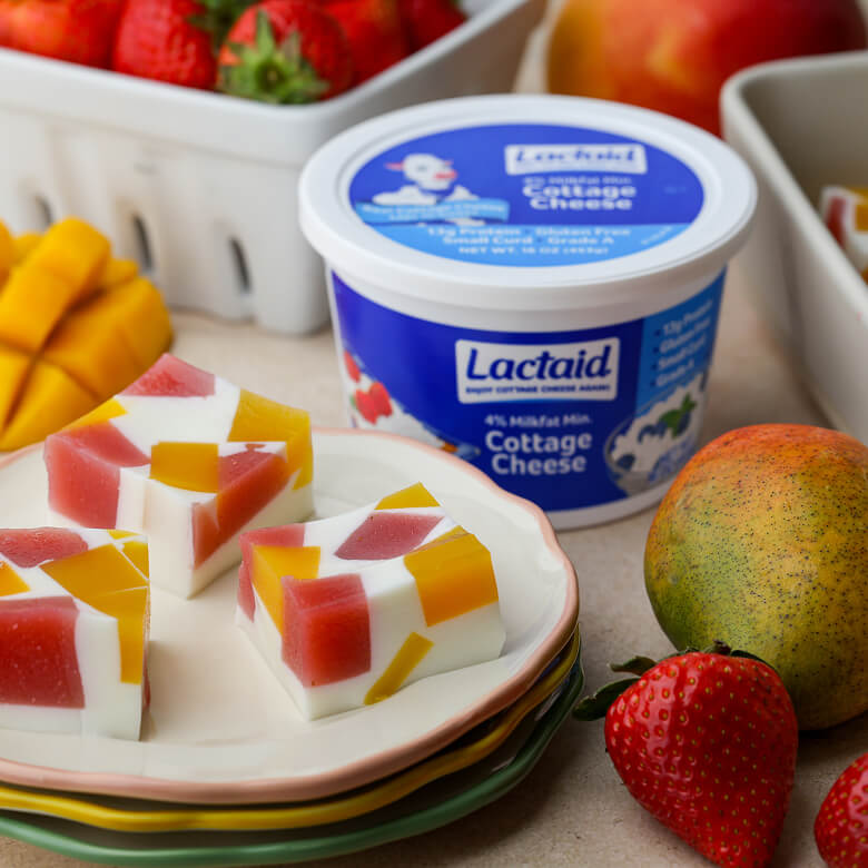 A container of LACTAID® Cottage Cheese, fresh fruit, Summer Jelly Cubes on a stack of plates, arranged on a table.