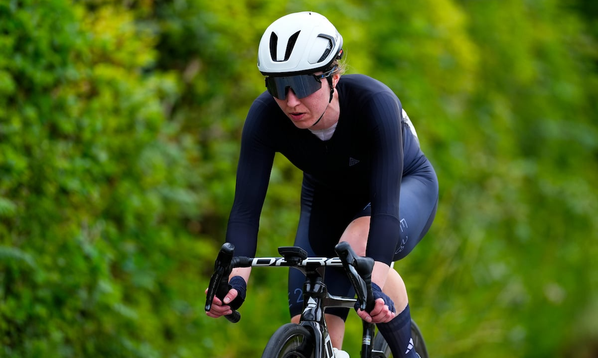 Anna rides a road bike wearing black kit, a white helmet and sunglasses against a blurred green leafy background.