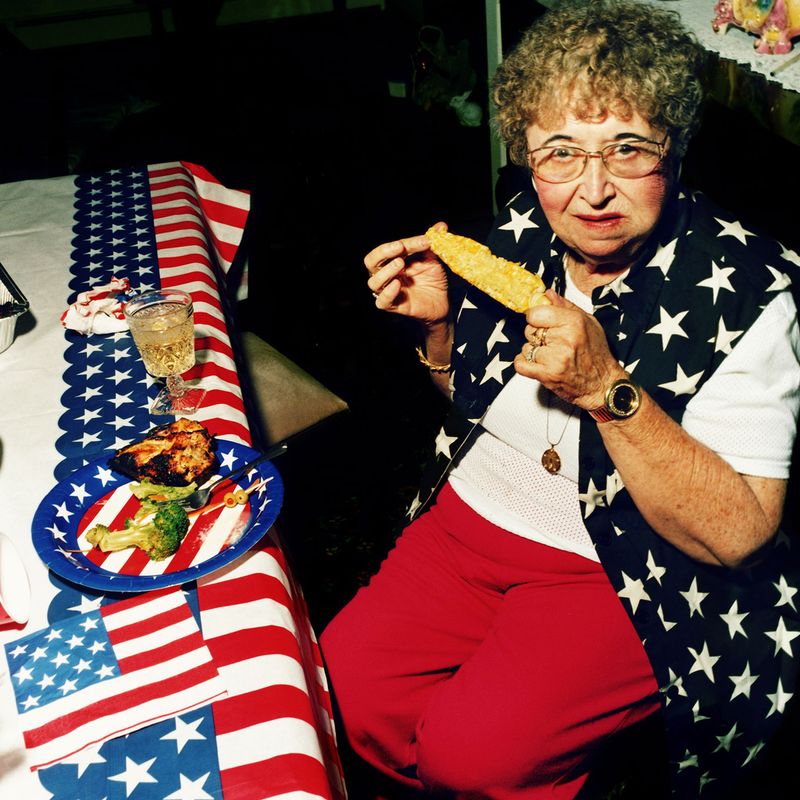 A photo of an older woman eating a corn on the cob while wearing American flag-themed clothes.