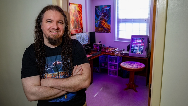 Terence Long poses with his arms crossed outside of his office, lit in purple with video games and Godzilla posters