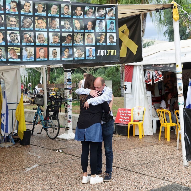 People embracing under a sign filled with photographs of Israeli hostages.