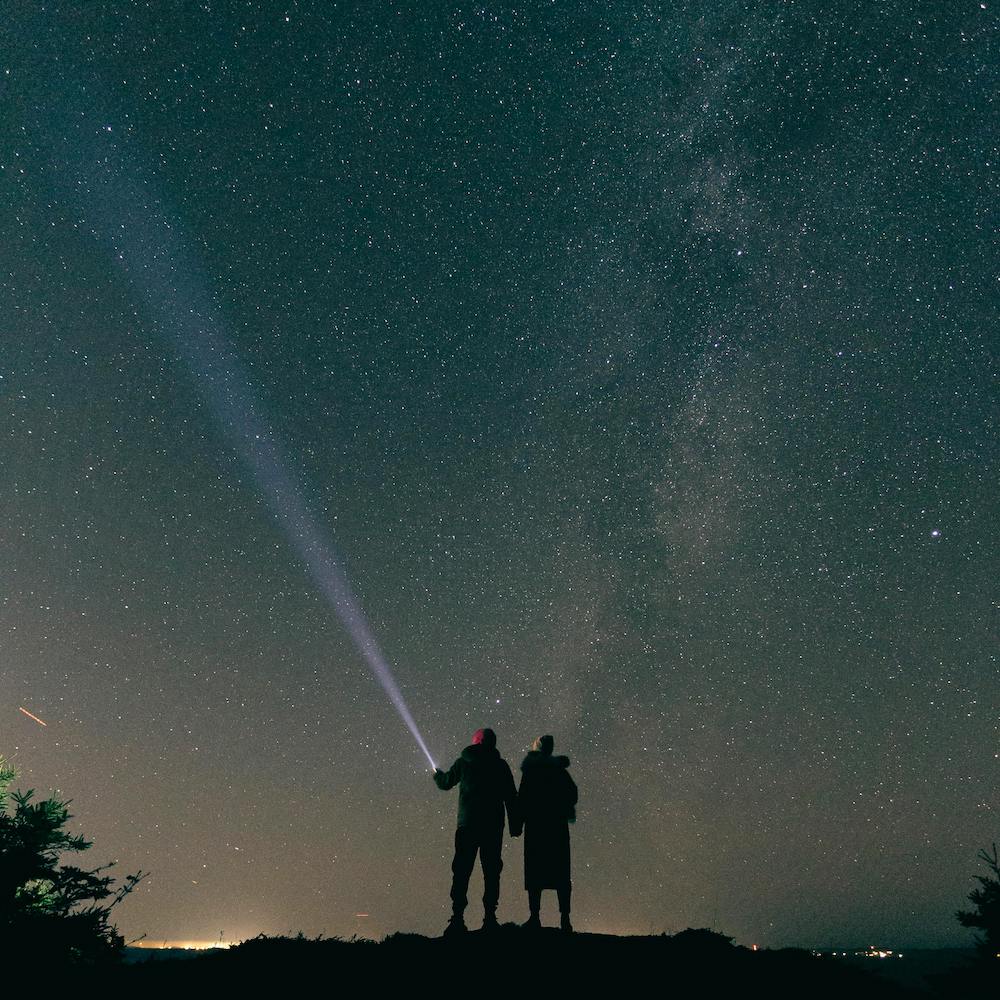 Two people stand in front of the night sky filled with stars, with the Milky Way visible. One of them is shining a flashlight into the sky.