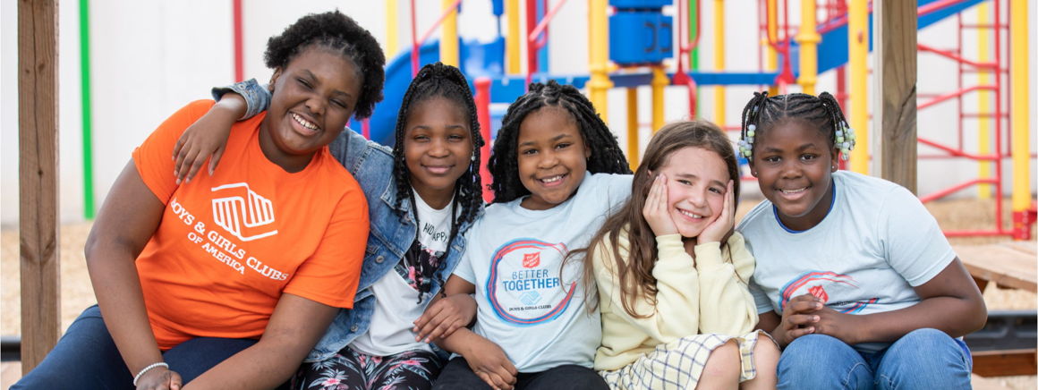 A group of five children and a caregiver smiling and sitting together on a playground. They wear colorful t-shirts, and the playground equipment is visible in the background.