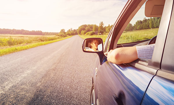 A blue car driving down a road at sunset. The driver’s arm is sticking out the open window.