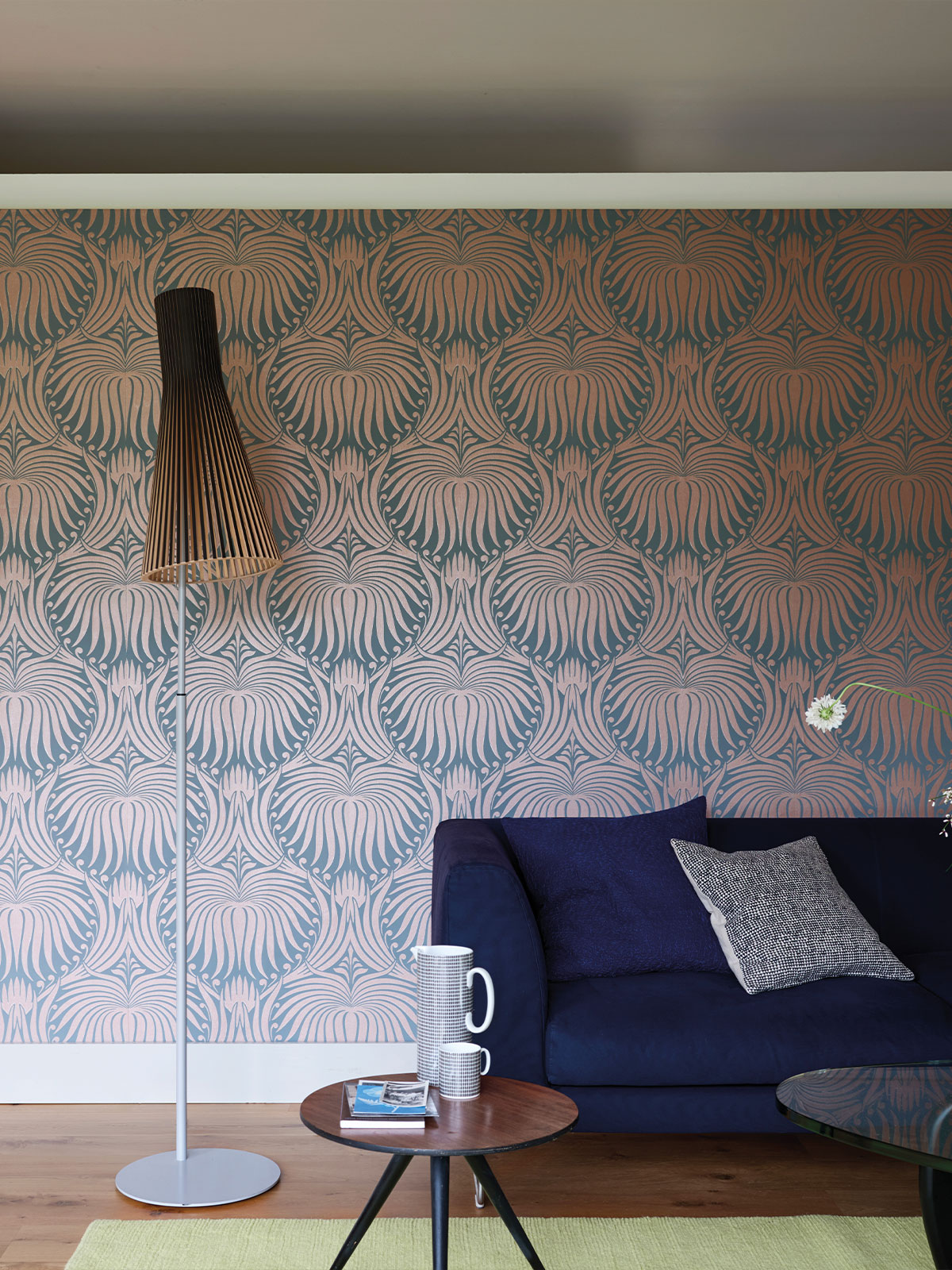 A modern living room with bold patterned wallpaper in copper and blue tones, a navy sofa with textured cushions, a slim white floor lamp with a wooden slatted shade, and a small round coffee table holding mugs and books on a wooden floor with a pale green rug.