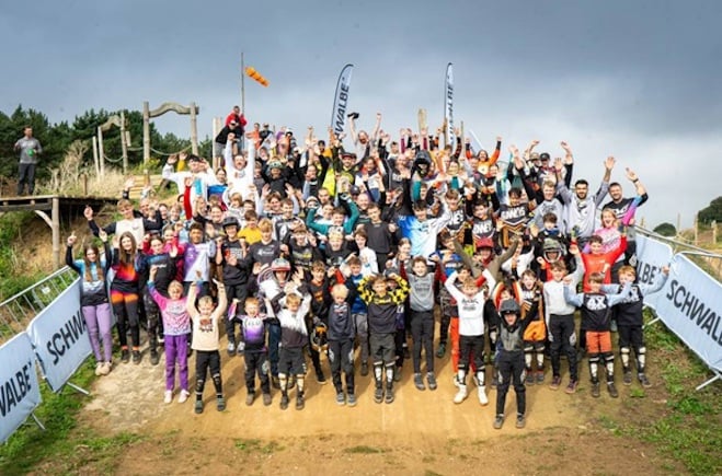 A big group of four cross riders pose for a photo on a dirt bank, with arms in the air.