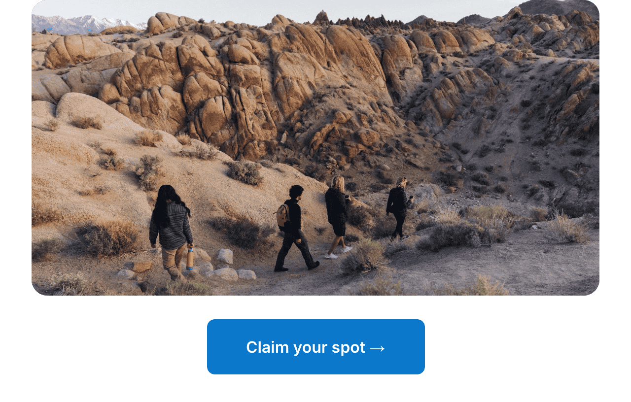 Four people hiking along a desert trail through rocky, boulder-strewn terrain with mountains in the background.