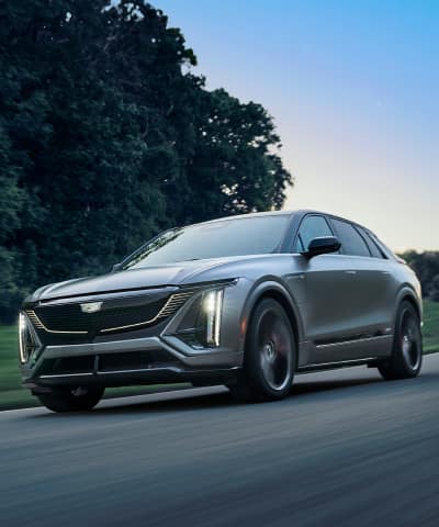 Cadillac LYRIQ-V driving down a road with trees, grass, and a clear blue sky in the background.