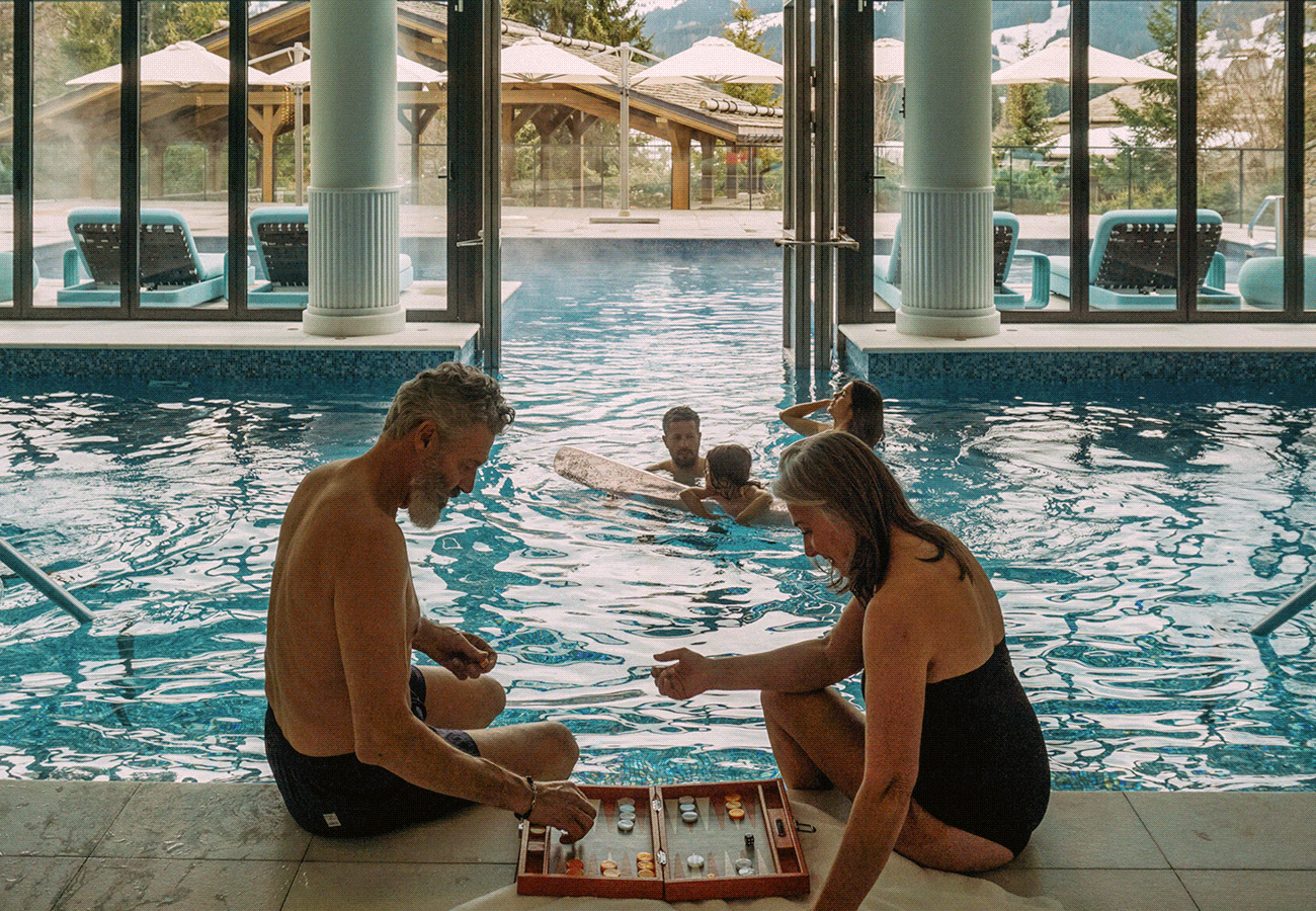 Hero GIF featuring 4 frames:  1) A couple playing a game beside a pool while a family swims nearby.  2) Children running behind a man carrying a torch, dressed in traditional Hawaiian attire.  3) A man standing on a balcony looking toward a large, snow-covered mountainside chalet. 4) Two women sharing small plates and wine on a rooftop lounge.