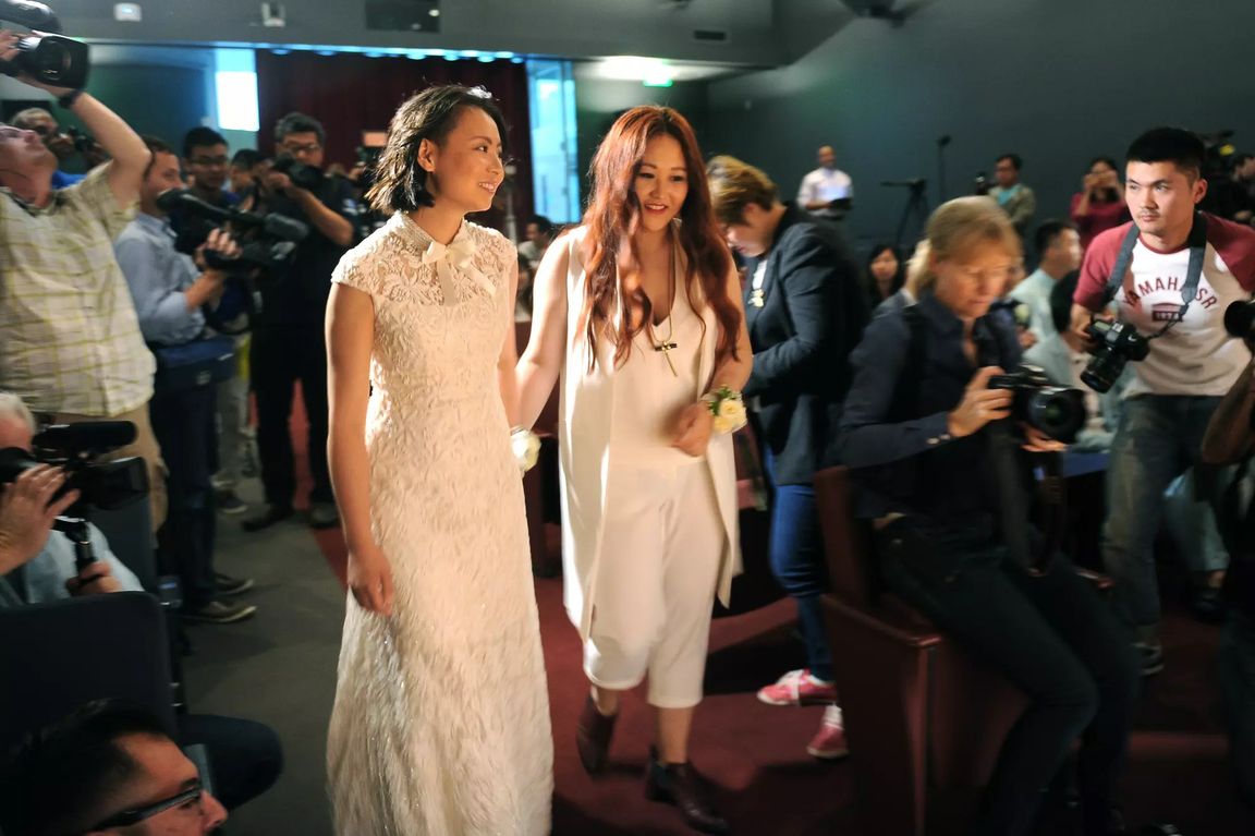 Two women in white hold hands in a room filled with photographers