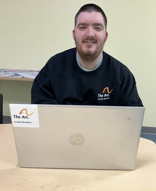Nick sits at a table with a laptop, smiling at the camera. He's wearing a The Arc Greater Brockton sweatshirt.