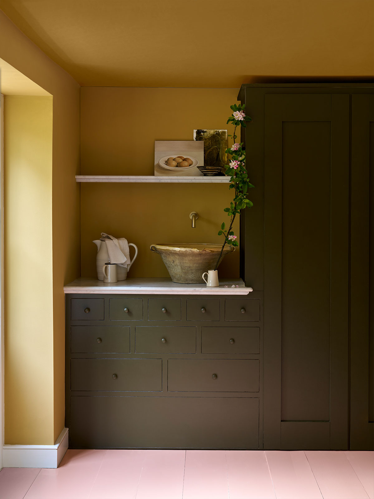 Diffused sunlight brightens this buttery yellow which blends into the moodier tone of the deep green wooden dresser. It gives the appearance of almost day and night, light and dark. The pink of the vine of blossom draping over the dresser picks up the tone of the floor, almost like the palest pink of the sky at sunrise.