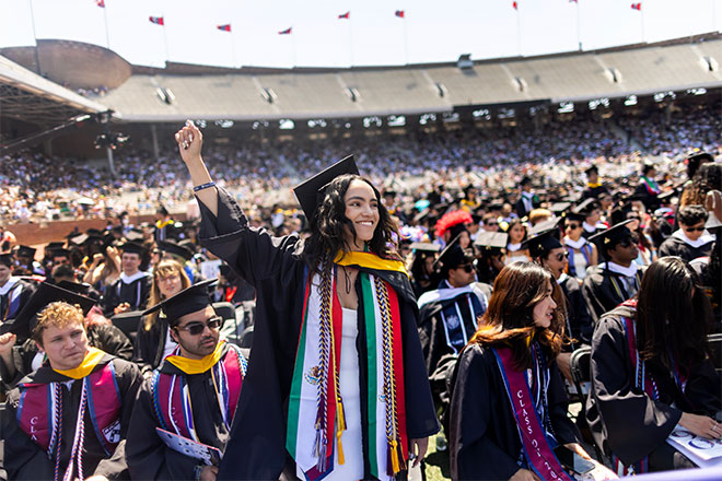 A graduating student stands up from a crowd, holding her fist up in the air.