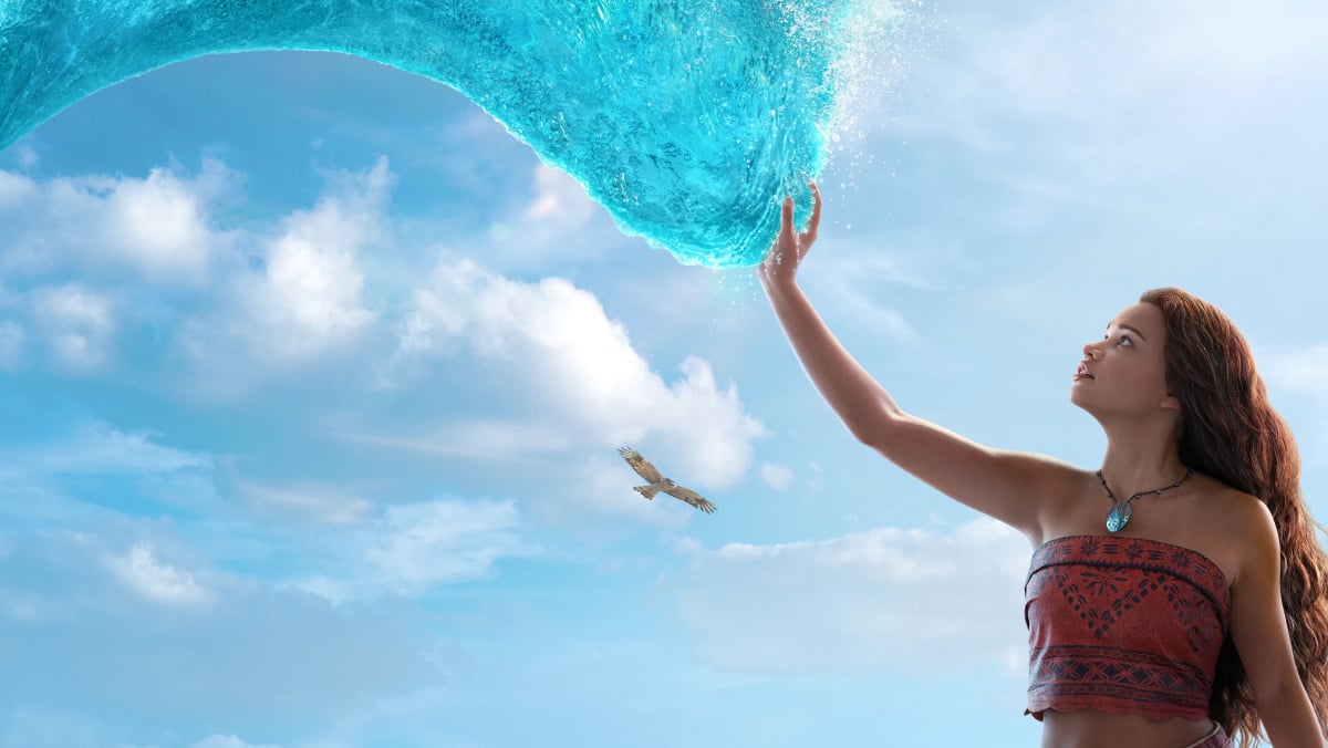 Moana stands underneath a cresting wave, gently touching it with her right hand. A large bird soars in the blue sky above her, as puffy white clouds drift by.