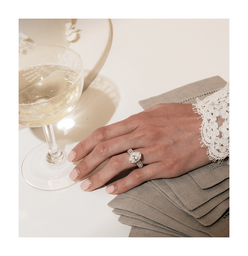 A hand wearing a ring with an oval cut diamond next to a glass of champagne. 