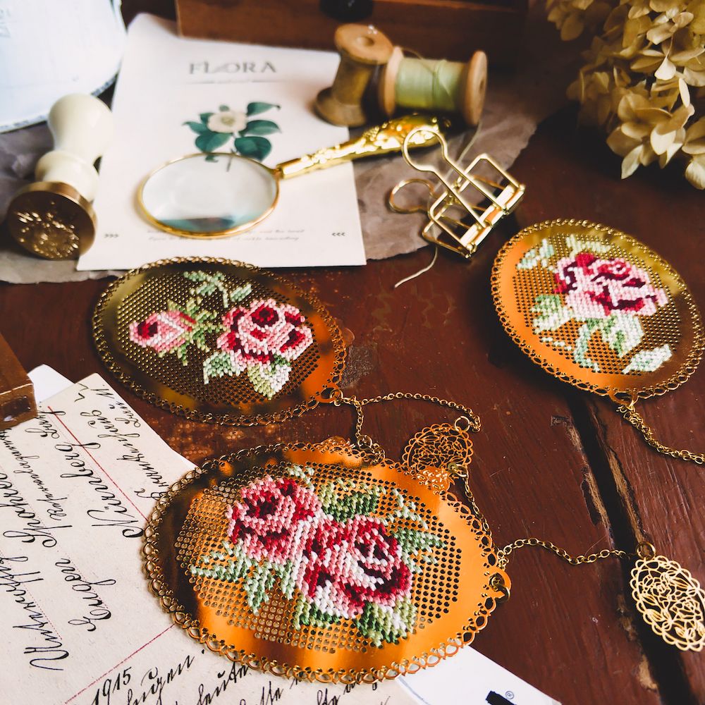A tabletop strewn with letters, spools of thread, a gold magnifying glass, and three gold ovals with roses cross-stitched on them. 
