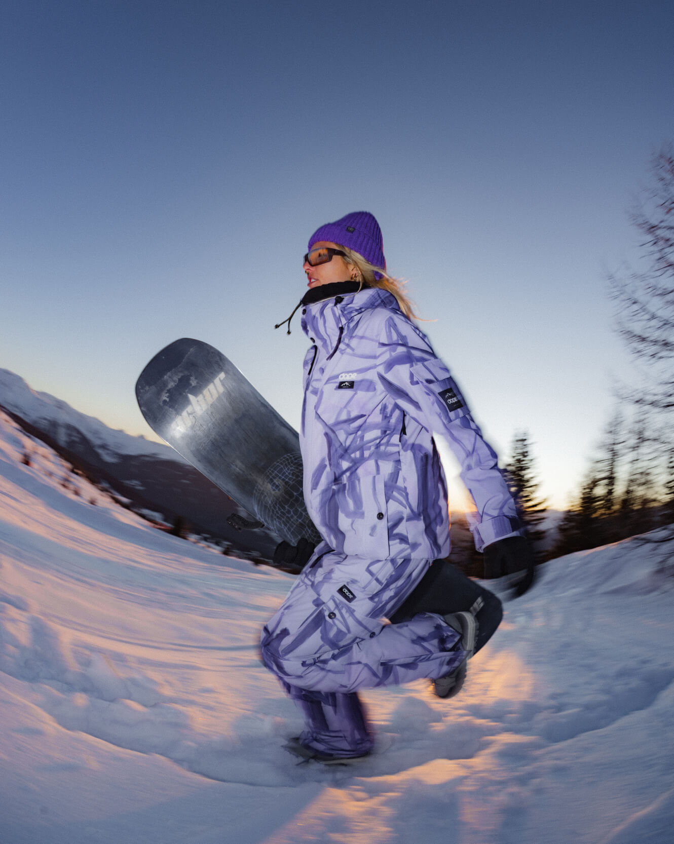 Woman in purple snowboard gear carrying board on snowy slope at sunset.