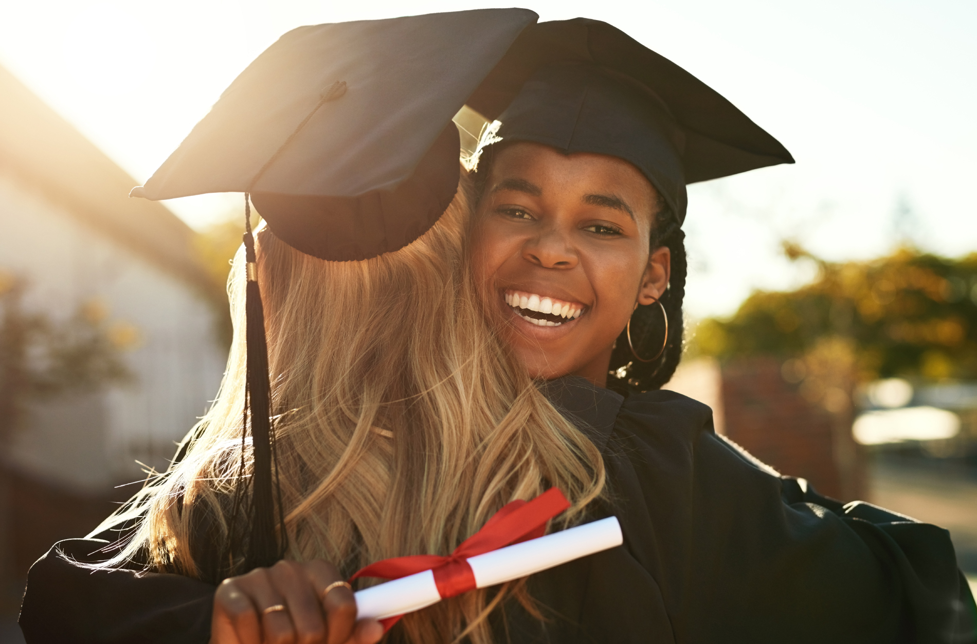 Smiling graduate in cap and gown holding diploma while hugging a friend outdoors at sunset