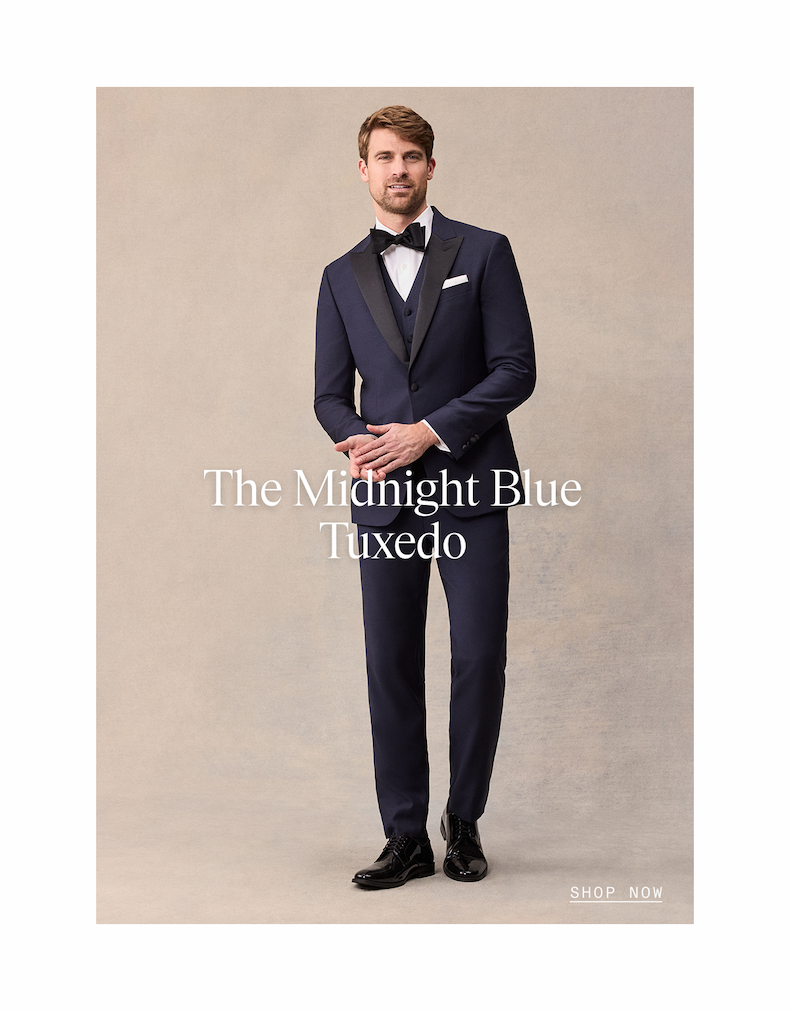 Man in a midnight blue three-piece tuxedo with black satin lapels, bow tie, and patent leather shoes, standing against a neutral backdrop. The Midnight Blue Tuxedo. SHOP NOW