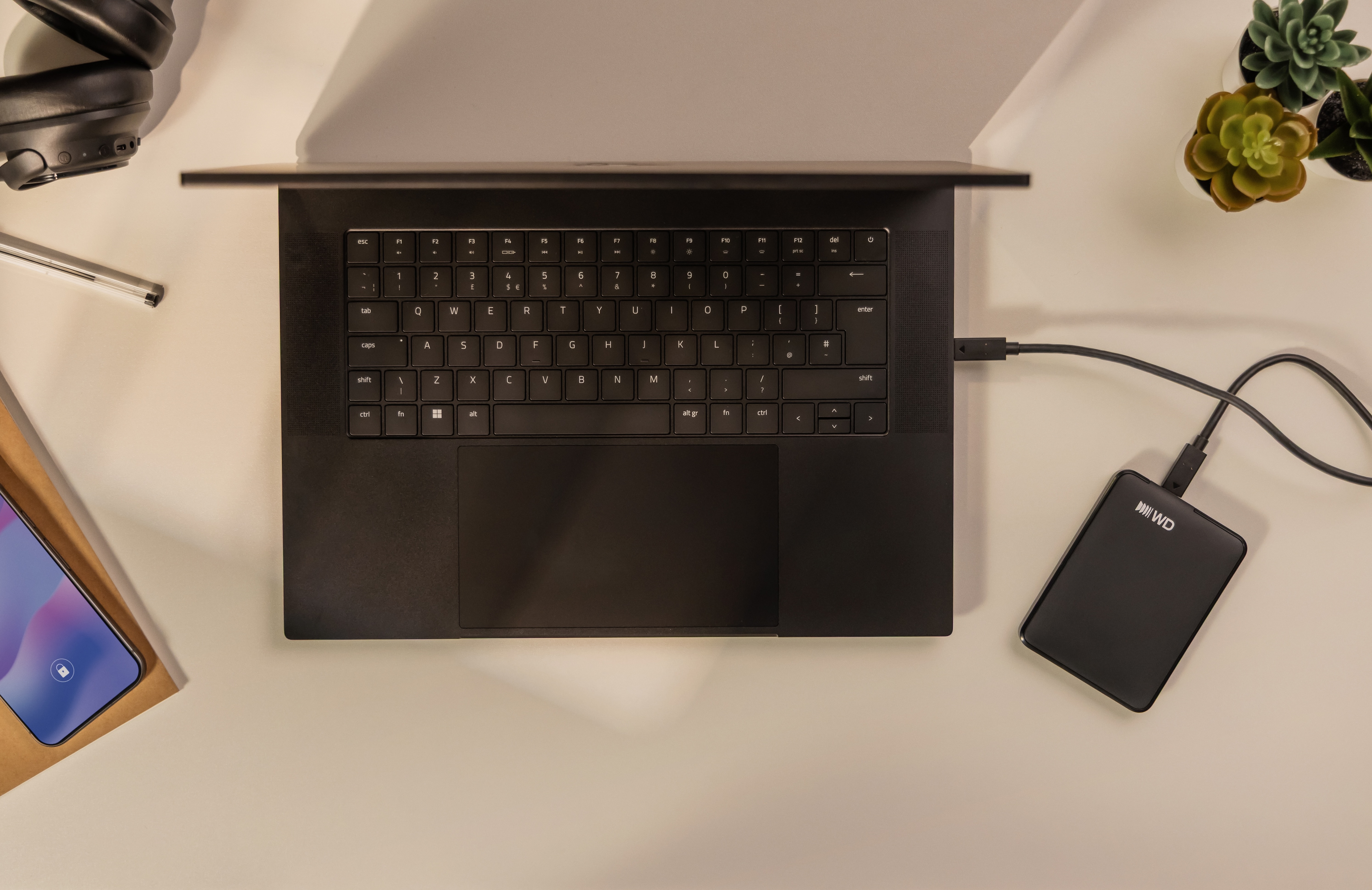 Overhead view of a desk featuring a black laptop, external hard drive, headphones, smartphone, and small potted plants.