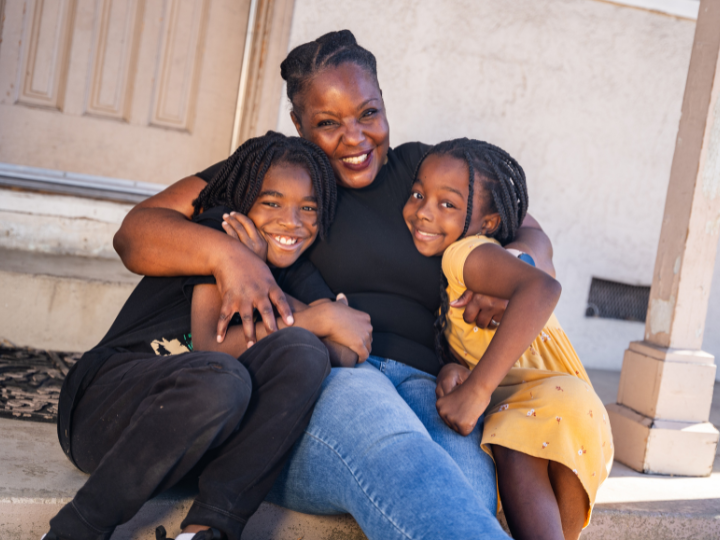 Zoe, mother of two, sits on the porch of her home with her two children, all three are smiling and hugging each other.