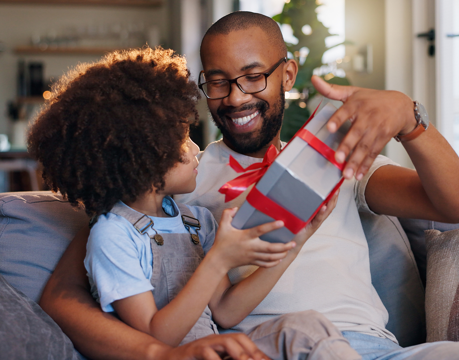 Smiling father wearing glasses opening a gift box with his young child while sitting together on a couch in a warmly lit living room.