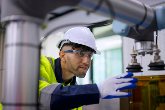 A man in a white hard hat, safety goggles and gloves examines a machine. 