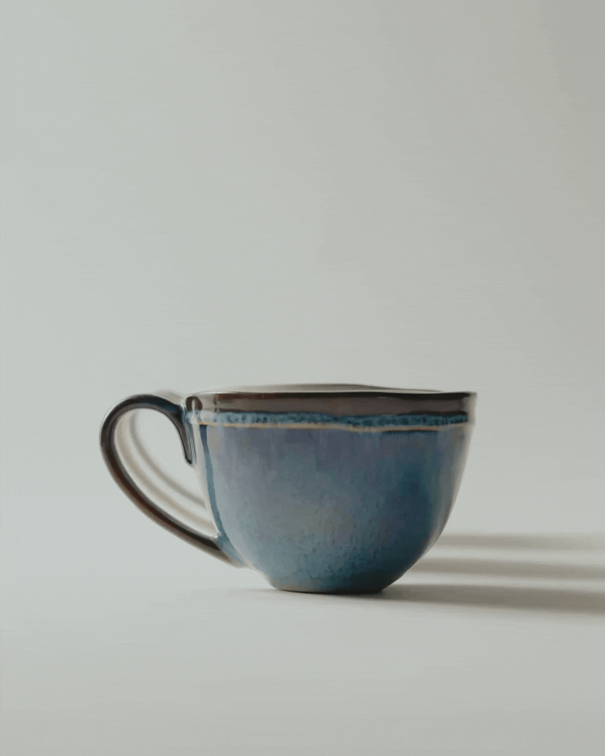 A simple blue ceramic cup with a curved handle, placed on a light background.