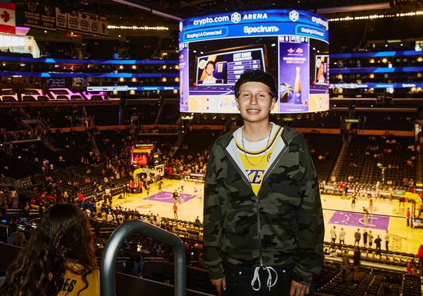 Young basketball player Ava Hernandez smiles courtside at a Lakers game at Crypto.com Arena, with the court and scoreboard visible below.