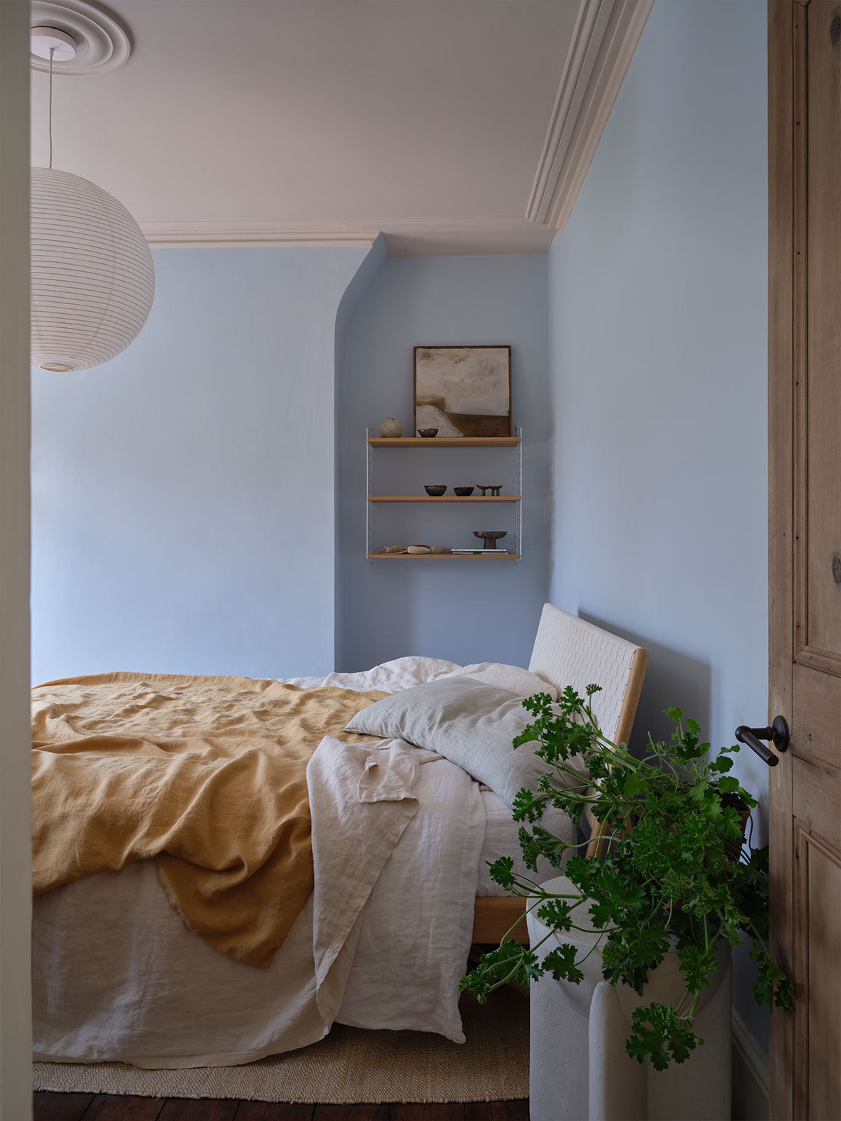 A bright and airy bedroom painted in pale blue, with layered linen bedding in warm neutrals and an ochre blanket. Minimalist shelves, natural textures and a leafy plant create a calm, soothing space filled with gentle light.