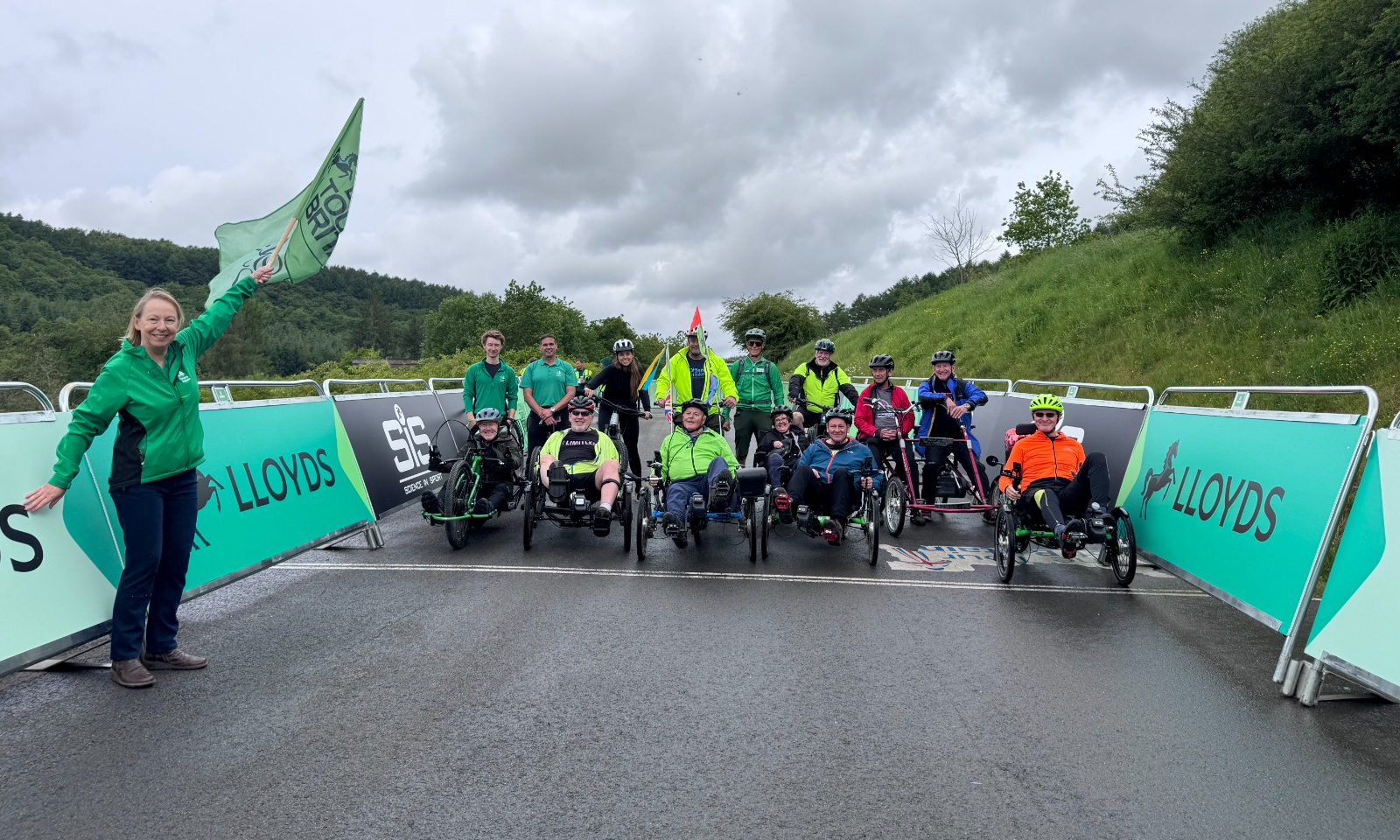 A group of riders on an assortments of bikes pause for a photo on the route of the Lloyds Tour of Britain, with a woman in the foreground waving a green flag.