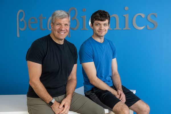 Photo: Beta Bionic’s Ed Damiano (left) with his son, David Damiano. A white man silver grey hair and wearing a black shirt and grey pants sits and poses next to a younger man with dark brown hair who is wearing a blue shirt and black pants. Both smile and pose in fornt of a blue
wall. Silver letters on wall read "Beta Bionic".