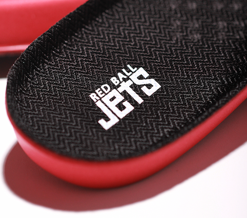 Close-up of a Red Ball Jets insole showing the textured black fabric top surface with a chevron weave pattern and embossed white Red Ball Jets logo. The red PU base of the insole is visible along the edge.