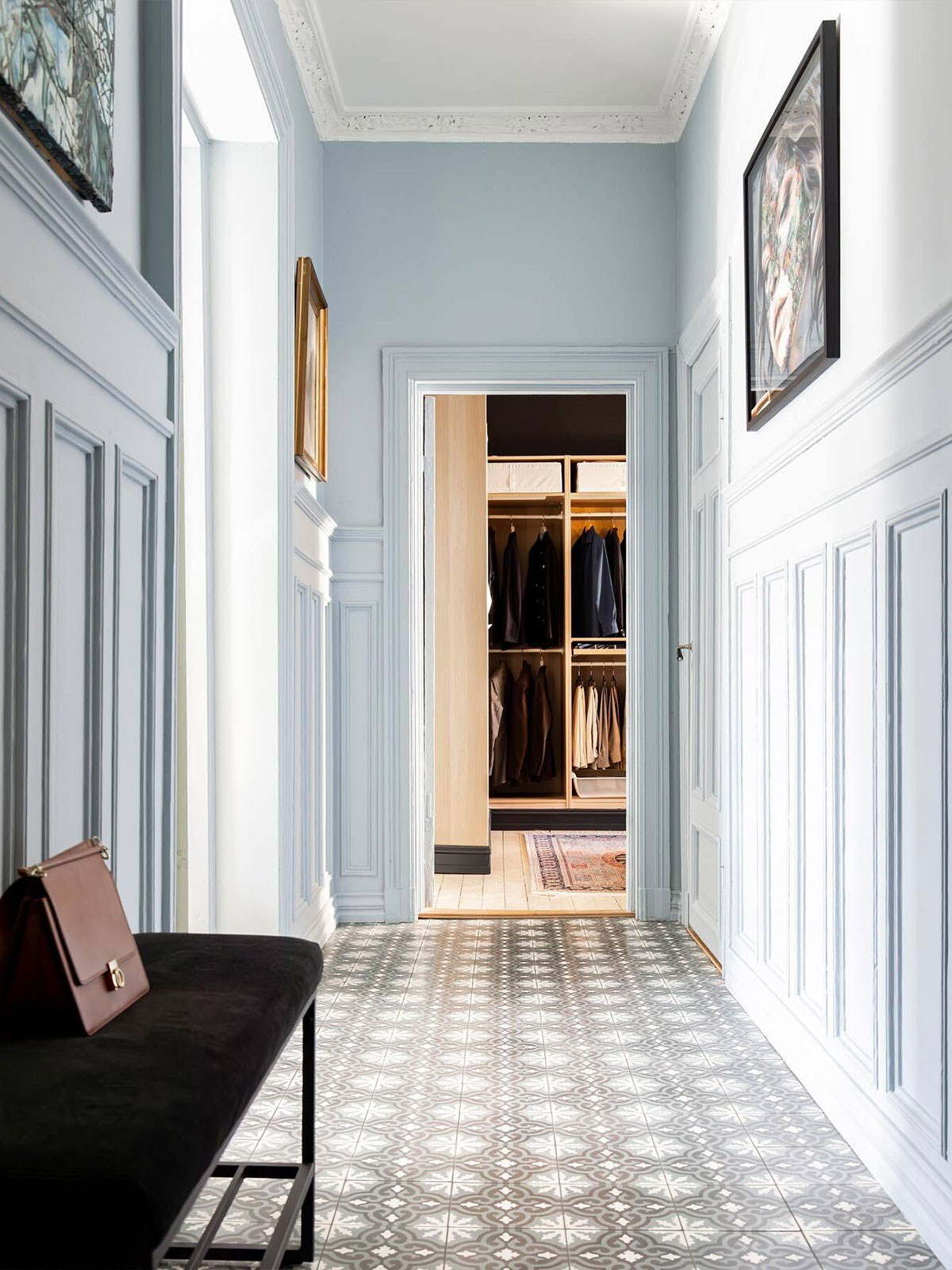 An elegant, panelled corridor leads to a well-organised dressing room beyond, with neat rows of jackets and trousers hanging in the wardrobe space. The corridor is painted in the palest blue, like the colour of an early summer sky. The brown briefcase on the black velvet bench suggests someone has just arrived home and is ready to relax.