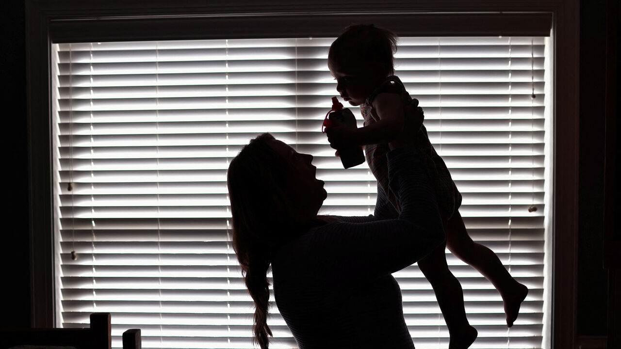 A woman holds up a toddler in Alabama USA. The image is almost black and white with a silhouette of the mother and child, with Venetian blinds in the background.