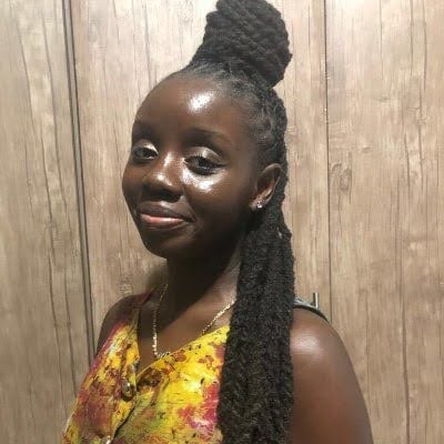 An African American woman wears her hair in locs in a hairstyle where her hair is in two-strand twists in a half-up, half-down style. She wears a tye-dye dress which is gold, pink, and green.