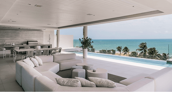 Open-plan living area with low seating and dining table beside a long infinity pool, overlooking palm trees and the ocean beyond.