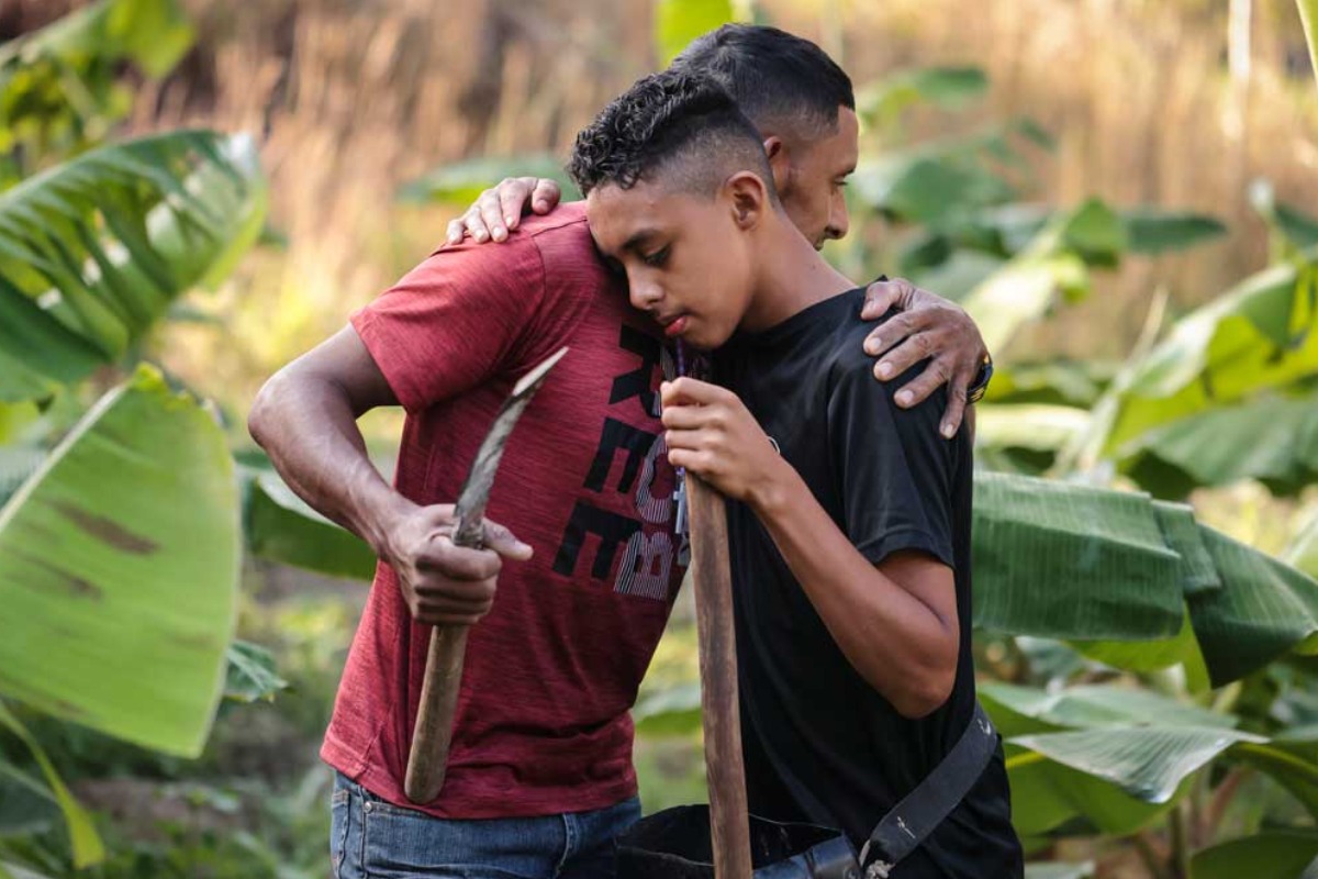 A man embraces his son as they stand together holding farming tools in a field of banana plants.