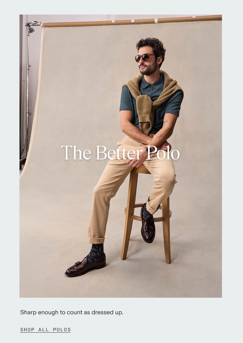 Man seated on a wooden stool in a studio setting, wearing a teal polo shirt, khaki chinos with cuffed hems, dark brown loafers, and patterned socks, with a tan knit sweater draped over his shoulders and round sunglasses. The Better Polo. Sharp enough to count as dressed up. SHOP ALL POLOS