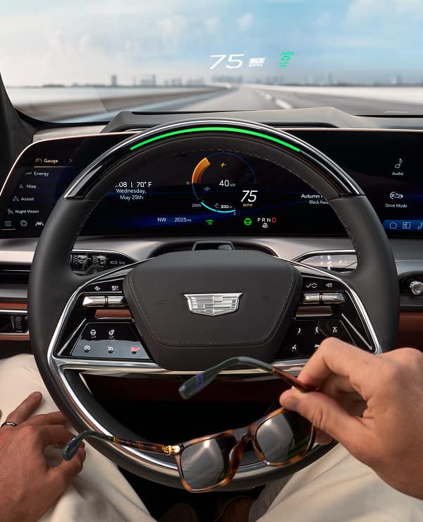 Front passenger seat view of the 2026 Cadillac VISTIQ’s steering wheel with Super Cruise activated; the driver is relaxed, looking ahead, and holding their sunglasses.