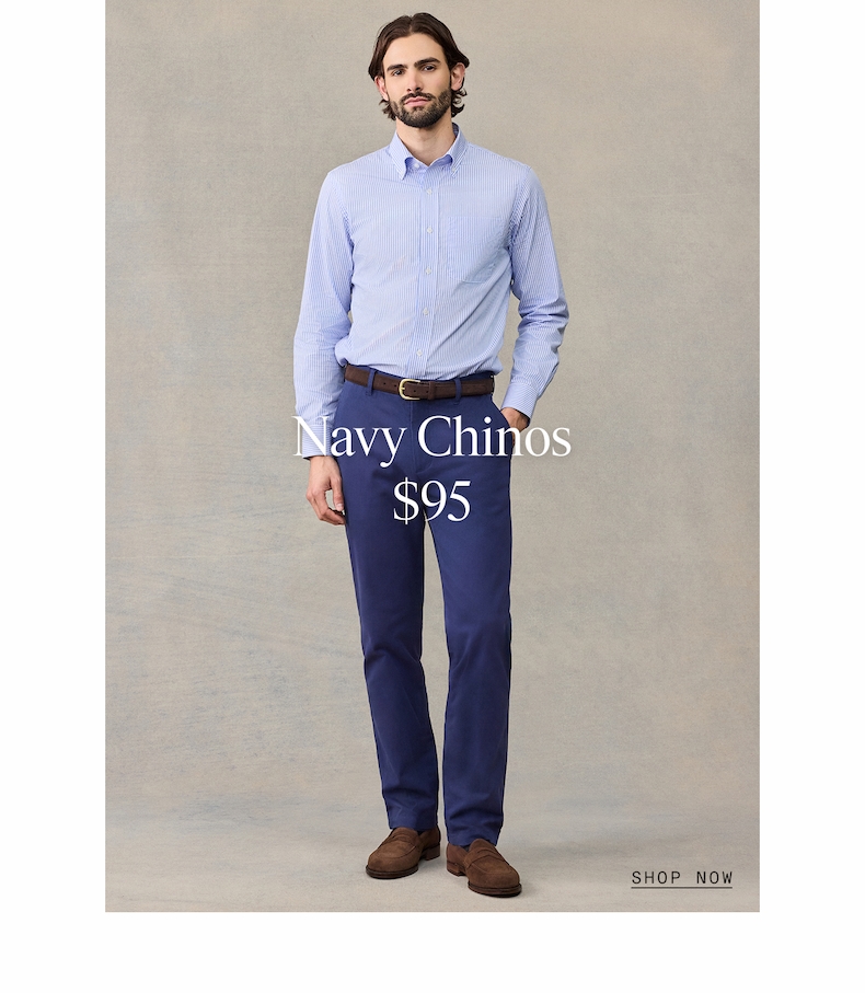 Full-length lifestyle photograph of a man wearing a light blue and white striped long-sleeve dress shirt, navy dress chinos, brown leather loafers, and brown belt, standing against a neutral beige background. Navy Chinos $95 SHOP NOW