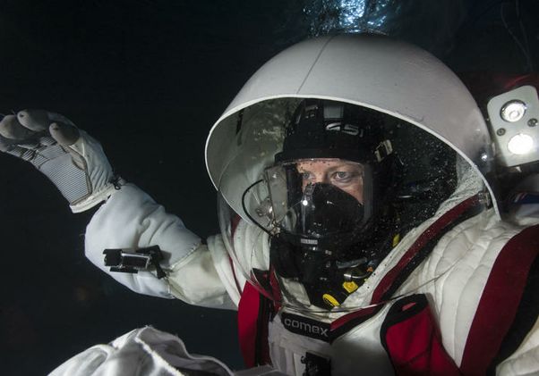 Woman in large white spacesuit, gloves and hood wears a breathing apparatus, holding her arms out in front