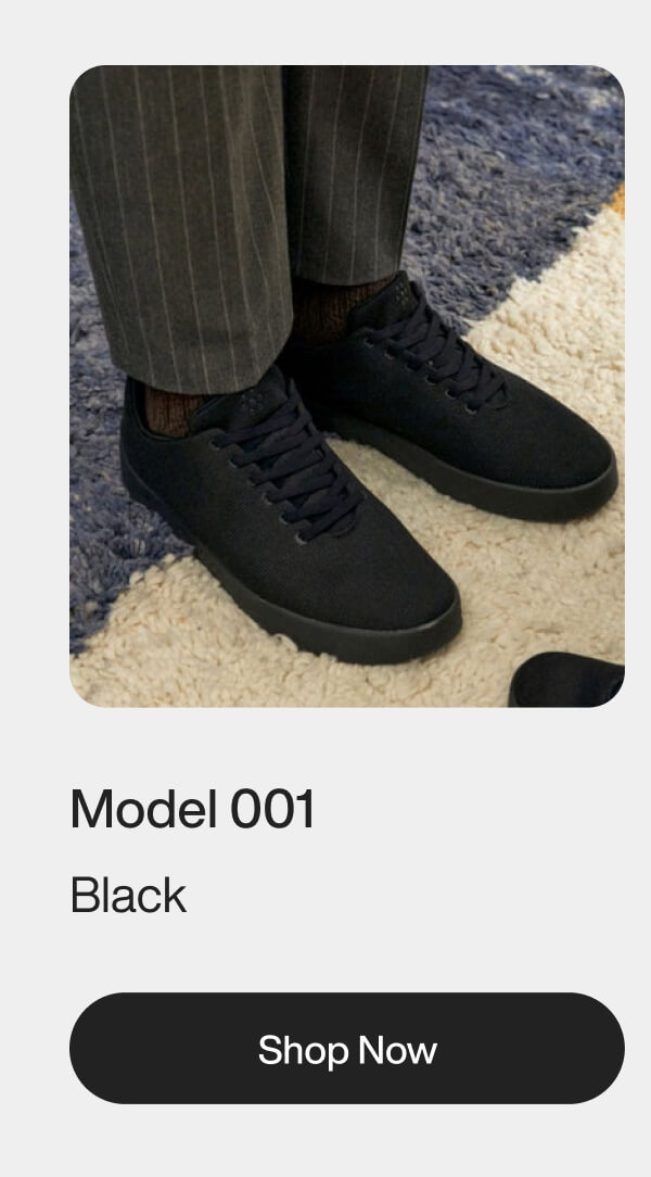 Man wearing black sneakers with pinstripe pants on a textured rug. Text reads "Model 001 Black" with a "Shop Now" button below.