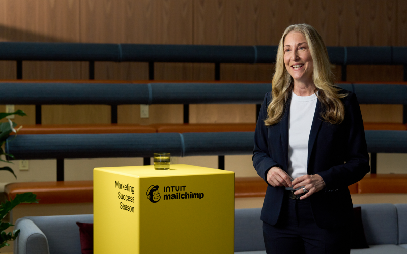 An icon of a megaphone, followed by the headline “Join the excitement.” Below, text reads “Unlock exclusive access to these fresh insights—all at no cost with your RSVP.” The time listed reads “12-1 pm UTC-4.” There is a call to action reading “Register now.”

To the right, speaker Tiffani Bova stands next to a yellow pedestal with the Intuit Mailchimp logo and a glass of water on it. She is wearing a navy blue blazer and a white shirt. 