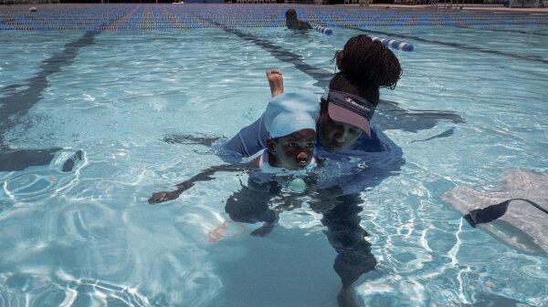 A woman wearing a visor hat and with her braids in a bun holds onto a young child wearing a blue swim cap in the pool. The woman is helping the child learn to swim. A ping pong ball is in front of the child in the water. 
