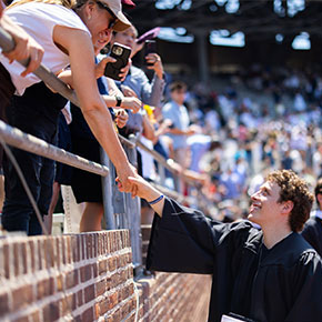 A student in a graduation robe reaches up to hold a family member's hand in the bleachers.
