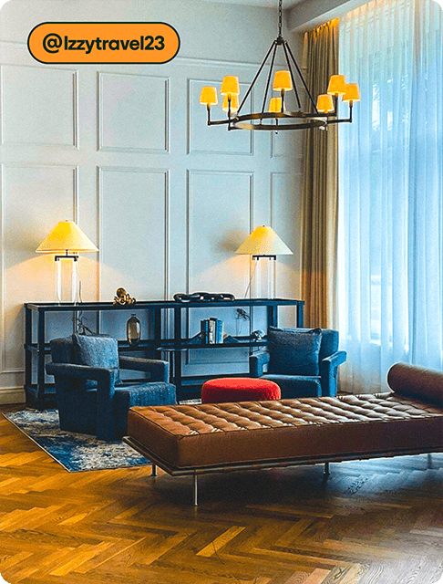 Image description: A sitting room at a hotel with white walls and a floor-to-ceiling window with sheer drapes. There’s a bookshelf with two lamps, two cozy chairs with a red ottoman between them, and brown leather chaise. A modern lighting fixture hangs above it all.
